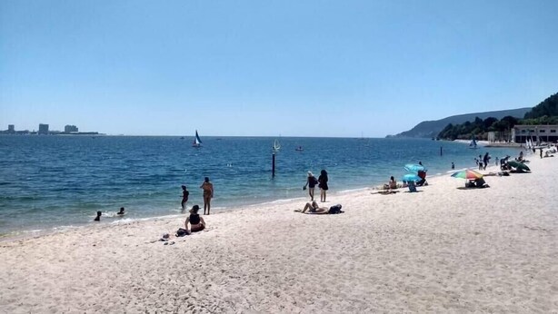 Une plage où des gens se prélassent au soleil et se baignent, un ciel dégagé et des bateaux sur l’eau.