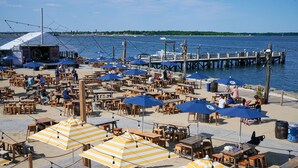 Un área de comedor al aire libre con mesas de madera y sombrillas azules, con vista a una masa de agua y un muelle al fondo.