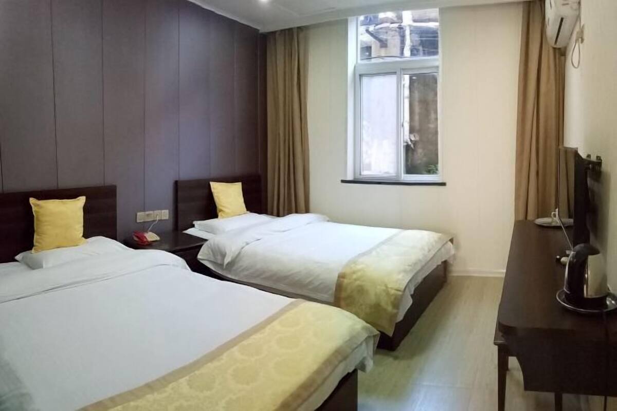 A hotel room with two beds, a desk, a window, and a wall-mounted air conditioner.