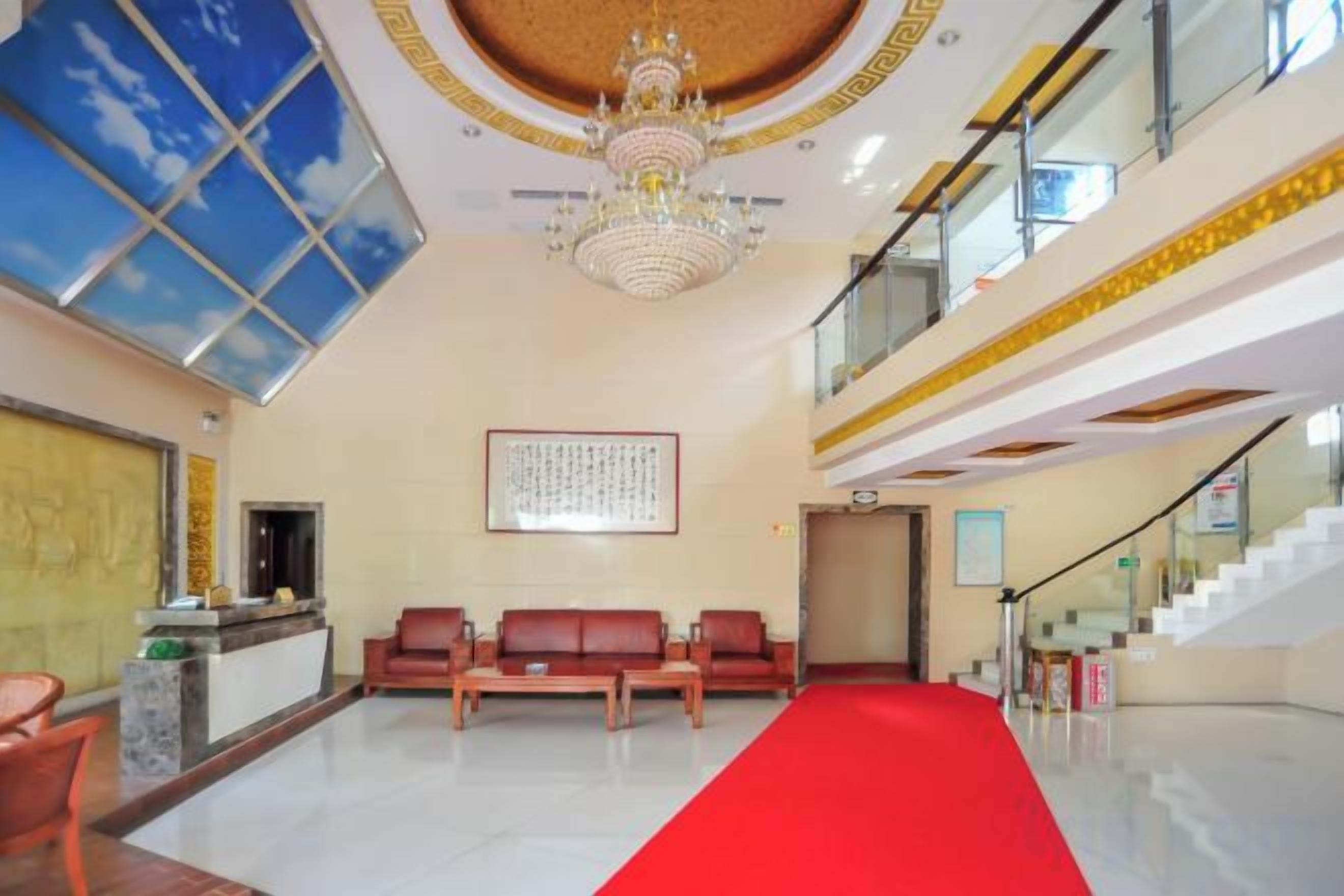 A hotel lobby with a red carpet, a reception desk, seating area, and a staircase.