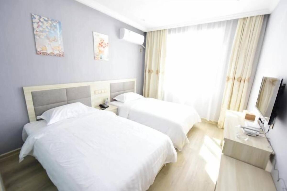 A hotel room with two beds, a desk, a chair, a flat-screen TV, and a window with curtains.