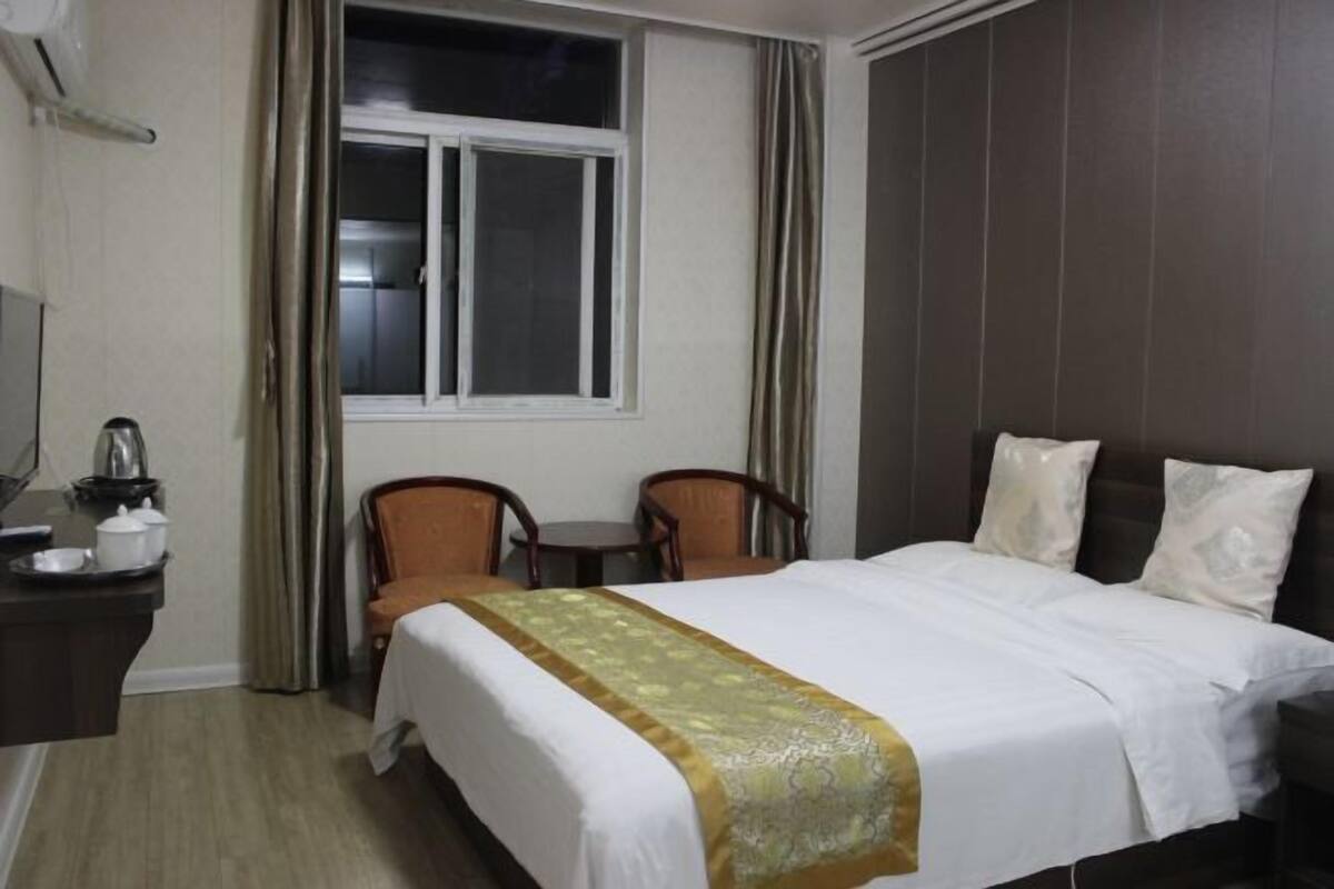 A hotel room with a bed, two chairs, a small table with a kettle, and a window with curtains.