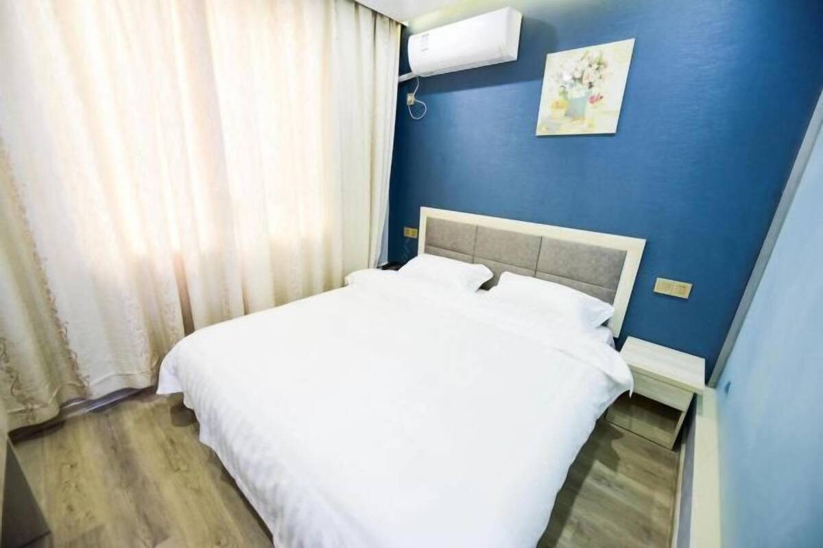 A hotel room with a bed, a nightstand, a wall-mounted air conditioner, and a painting on the wall.