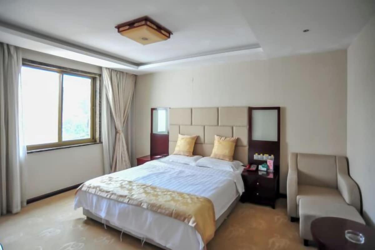 Una habitación de hotel con una cama grande, una mesita de noche con lámpara, una silla y una ventana con cortinas.