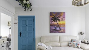 A room with a blue door, a beige sofa, a wall-mounted painting of a sunset, and a hanging plant. - Cosy and Bright in La Arena Beach (Zierbena)