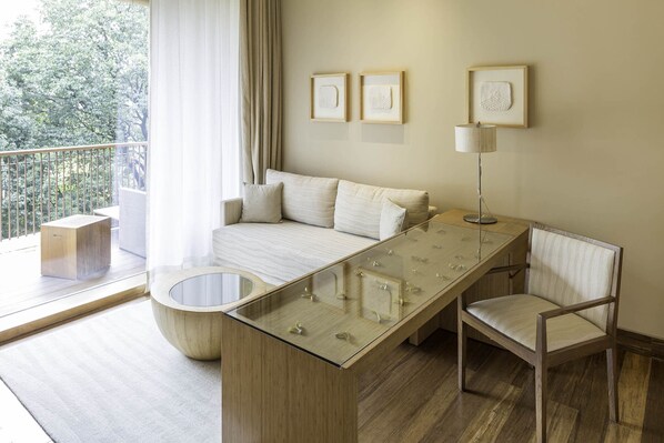 A modern living room with a glass coffee table, a sofa, a chair, and a floor lamp. There are framed artworks on the wall and a view of trees through the window.