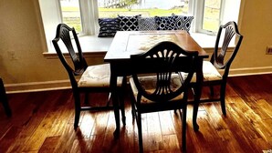 A dining area with a table and four chairs, a bay window with a view of a lake and dock, and a wooden floor. - Stunning Water Front Home with Private Dock and Beach and Sunsets (Lusby)