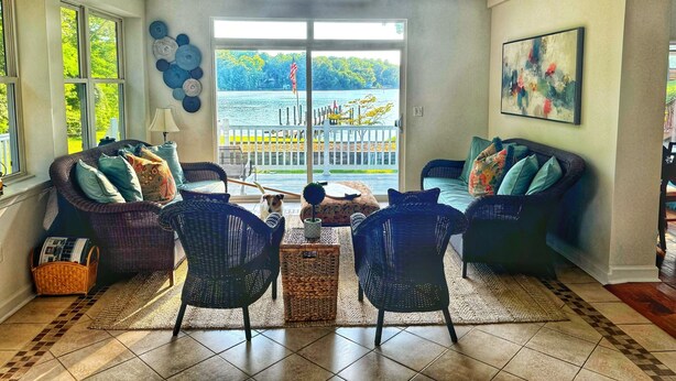 A spacious living room with a view of a lake, furnished with wicker chairs and a small table. - Stunning Water Front Home with Private Dock and Beach and Sunsets (Lusby)