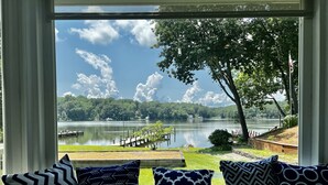 A view from a window with four patterned pillows on a windowsill, overlooking a lake with docks and boats, surrounded by greenery and trees. - Stunning Water Front Home with Private Dock and Beach and Sunsets (Lusby)