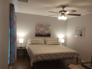 A bedroom with a bed, two bedside lamps, a ceiling fan, a painting on the wall, and a window with curtains.
