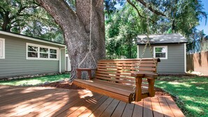 A wooden deck with a bench under a large tree, surrounded by a grassy yard and a small shed.