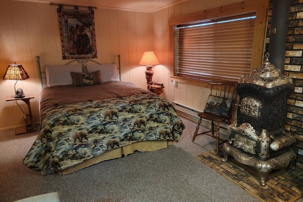A cozy bedroom with a bed, two lamps, a window with blinds, and a decorative stove.