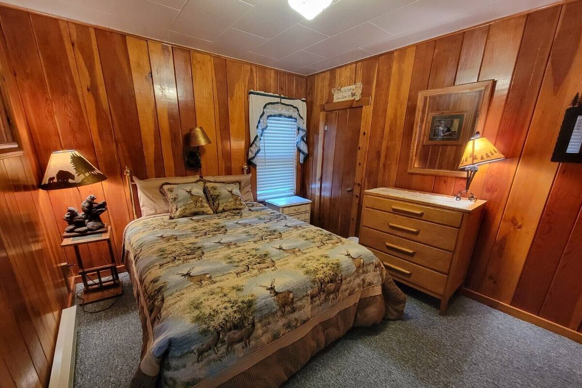 A wooden-paneled bedroom with a bed, nightstands, a dresser, and a window with blinds.