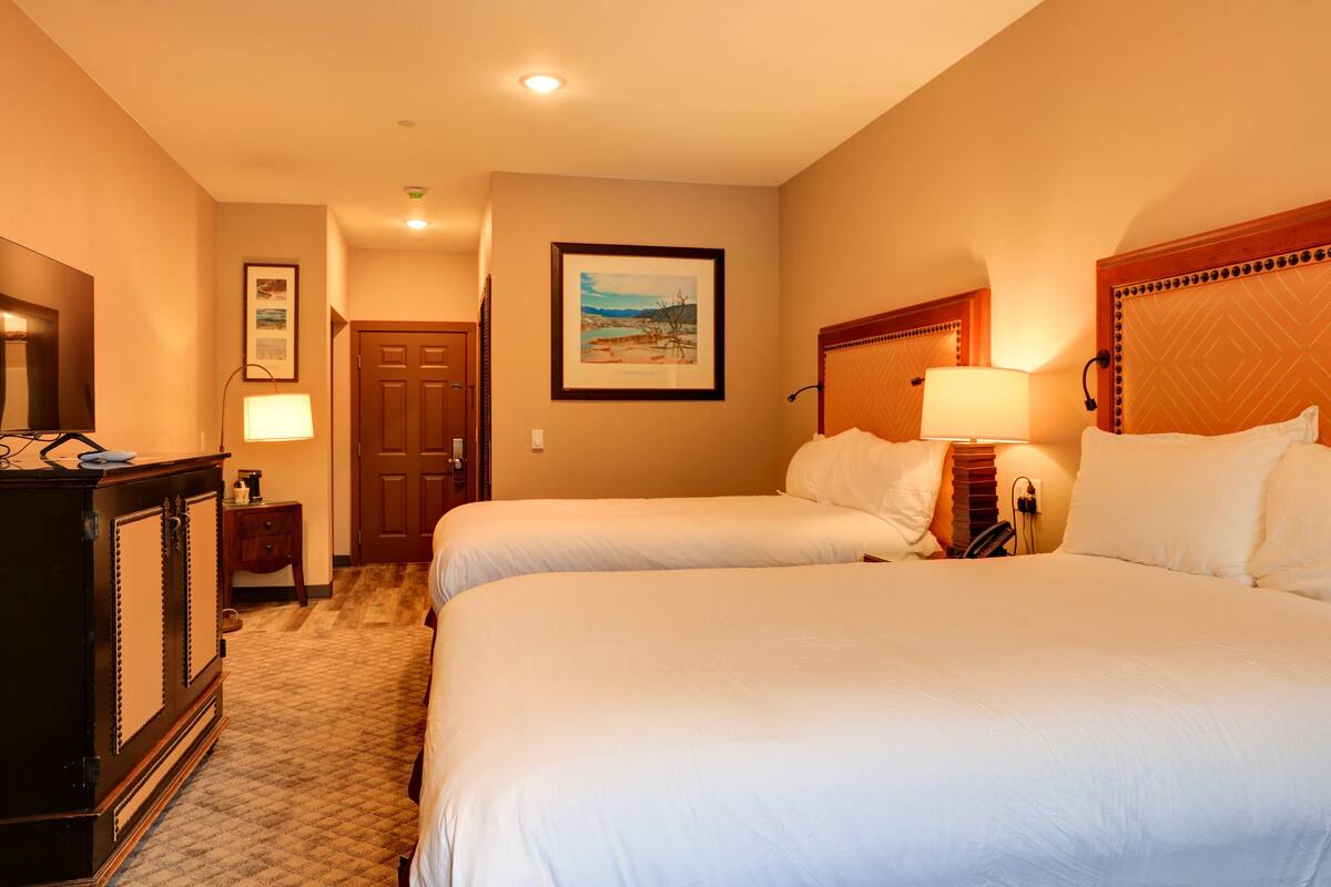 A hotel room with two beds, a wooden headboard, a nightstand with a lamp, a dresser, and a framed picture on the wall.