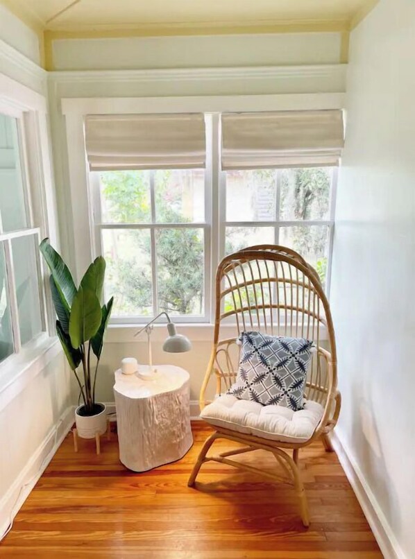 Habitación de esquina con piso de madera, una silla de mimbre con cojín estampado, una pequeña mesa redonda con lámpara y una planta en maceta.