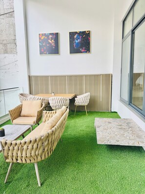 Een moderne zithoek met een bank, stoelen en een bijzettafeltje, met een vloer van kunstgras en twee abstracte schilderijen aan de muur.