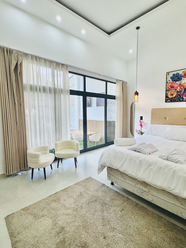 A modern bedroom with a large bed, a seating area with two chairs, a large window with curtains, and a painting on the wall.