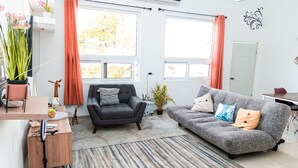 Sala de estar con sofá gris, un sillón negro y un tapete a rayas. Hay dos ventanas con cortinas, una planta en maceta y una mesita con un florero.