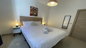 Una cama bien hecha con sábanas blancas, dos lámparas de noche, un cabecero, un cuadro abstracto colorido en la pared y una puerta de madera.
