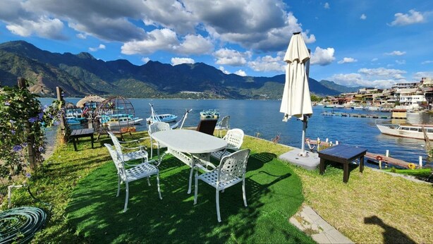 Un patio junto al lago con una mesa y sillas, barcos y montañas al fondo.