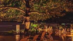 Un área de estar al aire libre con sillas y mesas bajo un árbol grande, rodeada de plantas y un letrero.