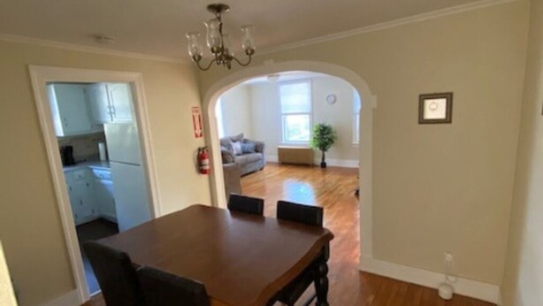 Uma sala de jantar com mesa e cadeiras de madeira, uma porta arqueada que dá para uma sala de estar com sofá e planta, e uma cozinha visível pela porta.