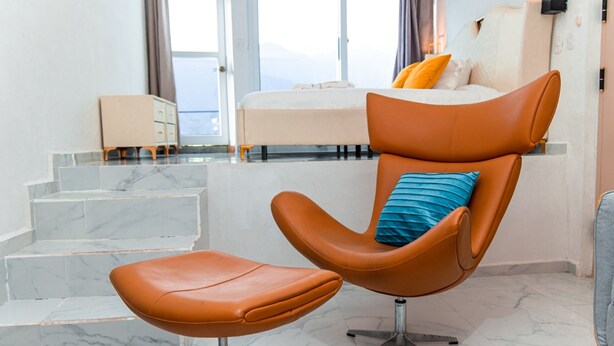 A modern hotel room with a curved leather chair, a small ottoman, a bed with orange pillows, and a view of the mountains through large windows.