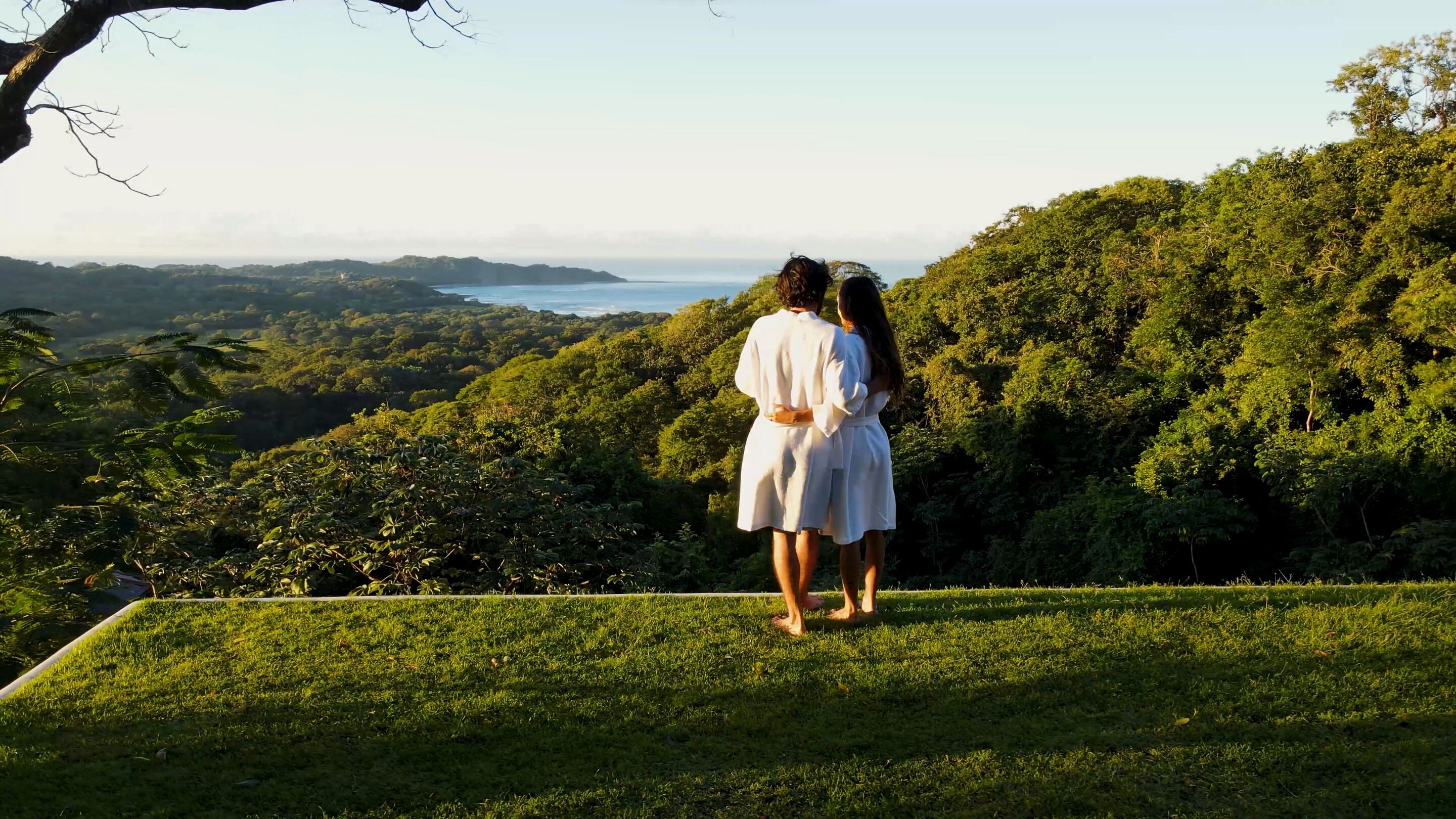 Two people standing on a grassy hill overlooking a lush landscape with a body of water in the distance.