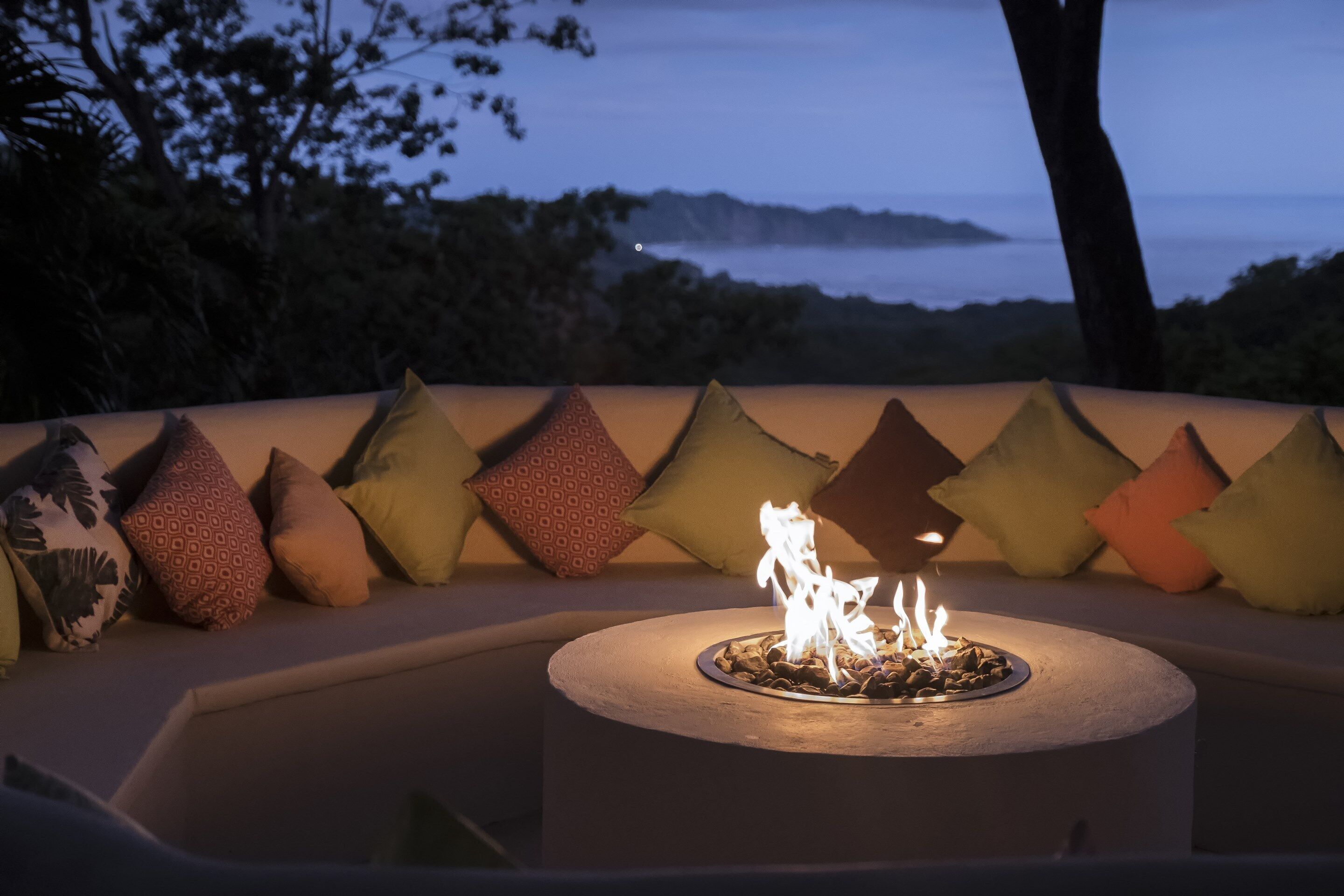A circular outdoor seating area with a fire feature, surrounded by a row of patterned cushions.