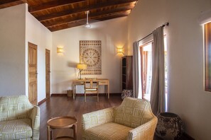 A spacious living area with a wooden ceiling, a desk with a chair, two patterned sofas, a round table, and a large decorative wall piece.