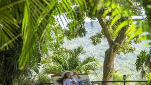 A person relaxing on a wooden deck chair by a pool, surrounded by lush greenery and a view of a mountainous landscape.
