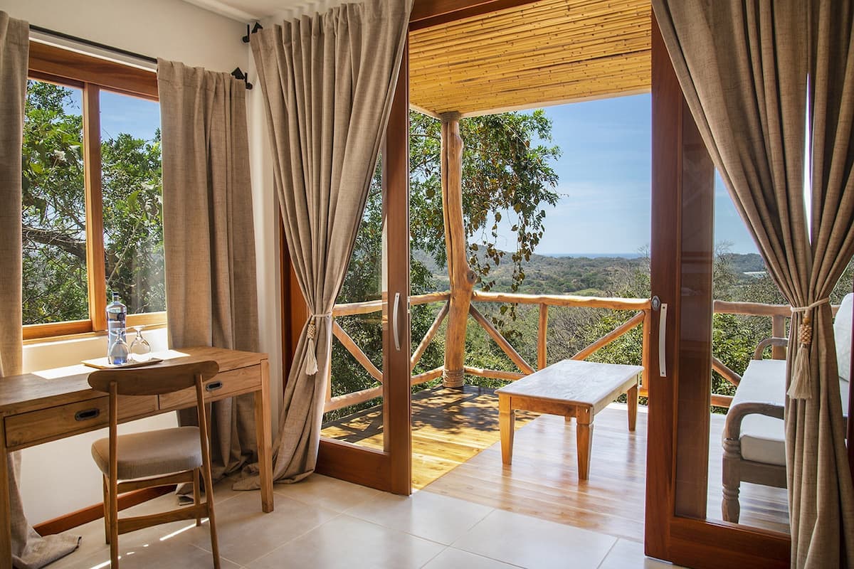 Ein Zimmer mit Holzschreibtisch, Stuhl und Bank, großen Fenstern mit Vorhängen und Blick auf eine gebirgige Landschaft.