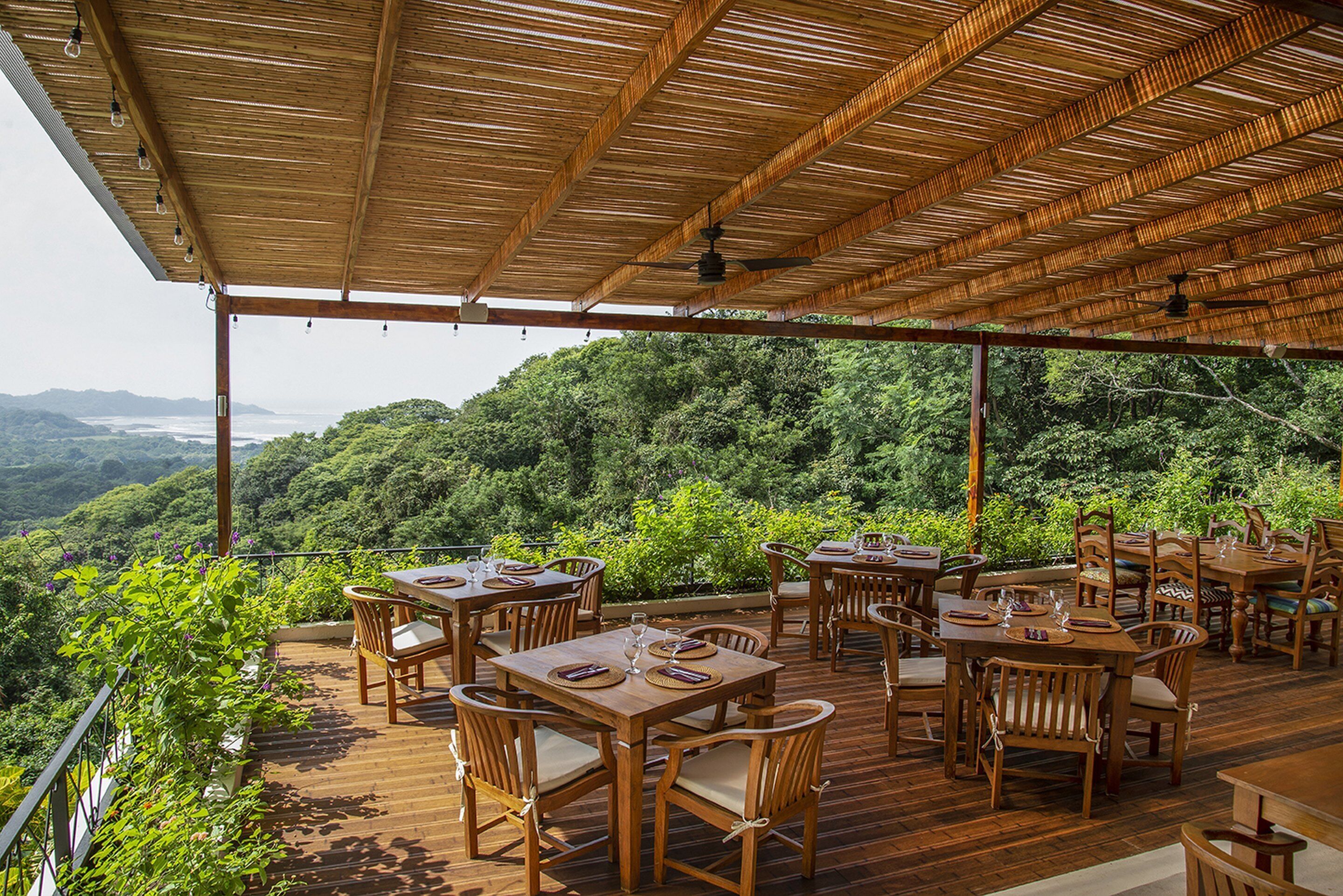 An outdoor dining area with wooden tables and chairs, surrounded by lush greenery and a view of the landscape.