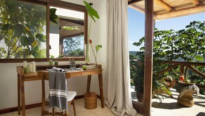 A wooden desk with a chair, a window with curtains, and a view of greenery outside.