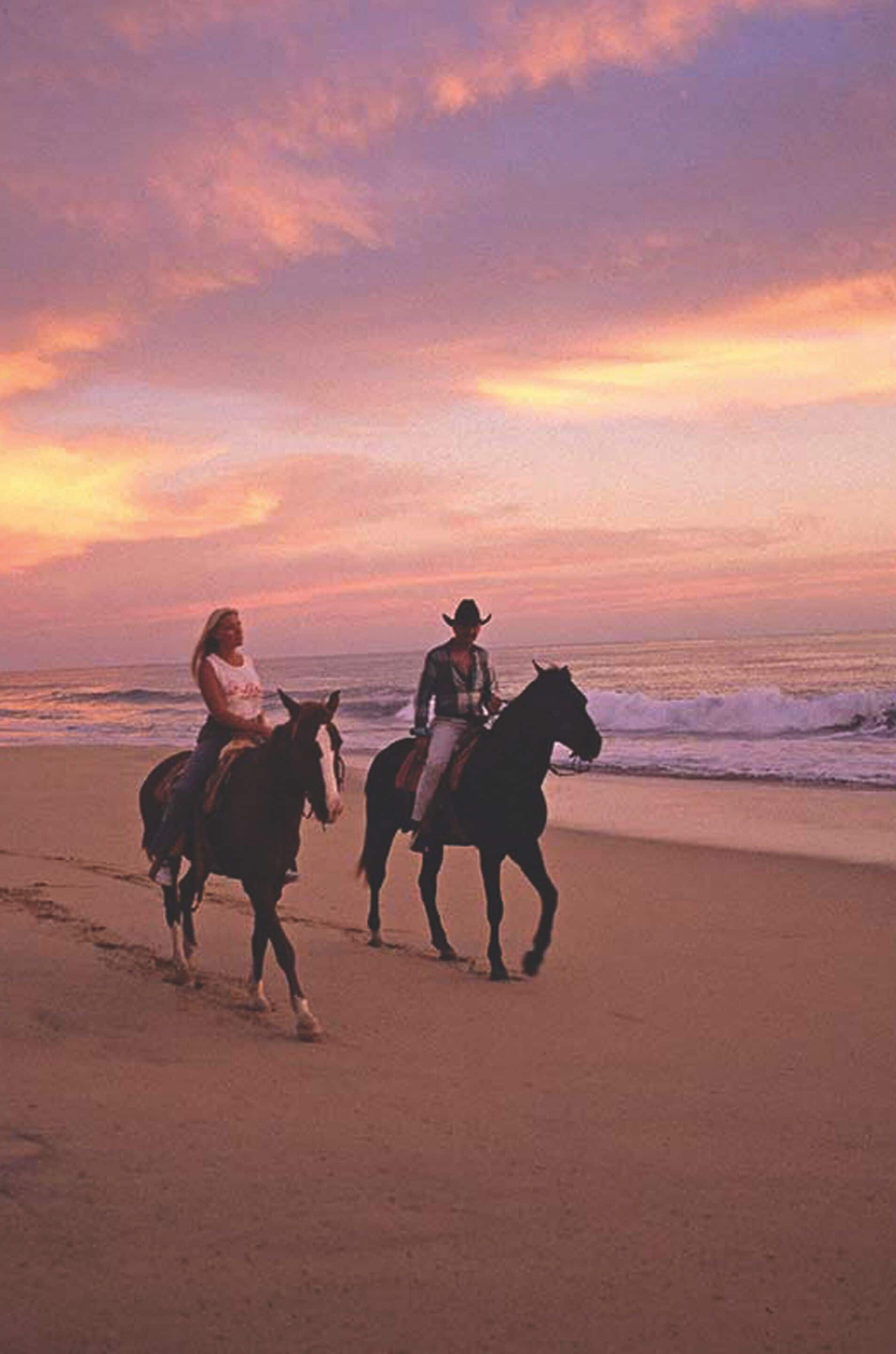 Zwei Personen reiten bei Sonnenuntergang am Strand.