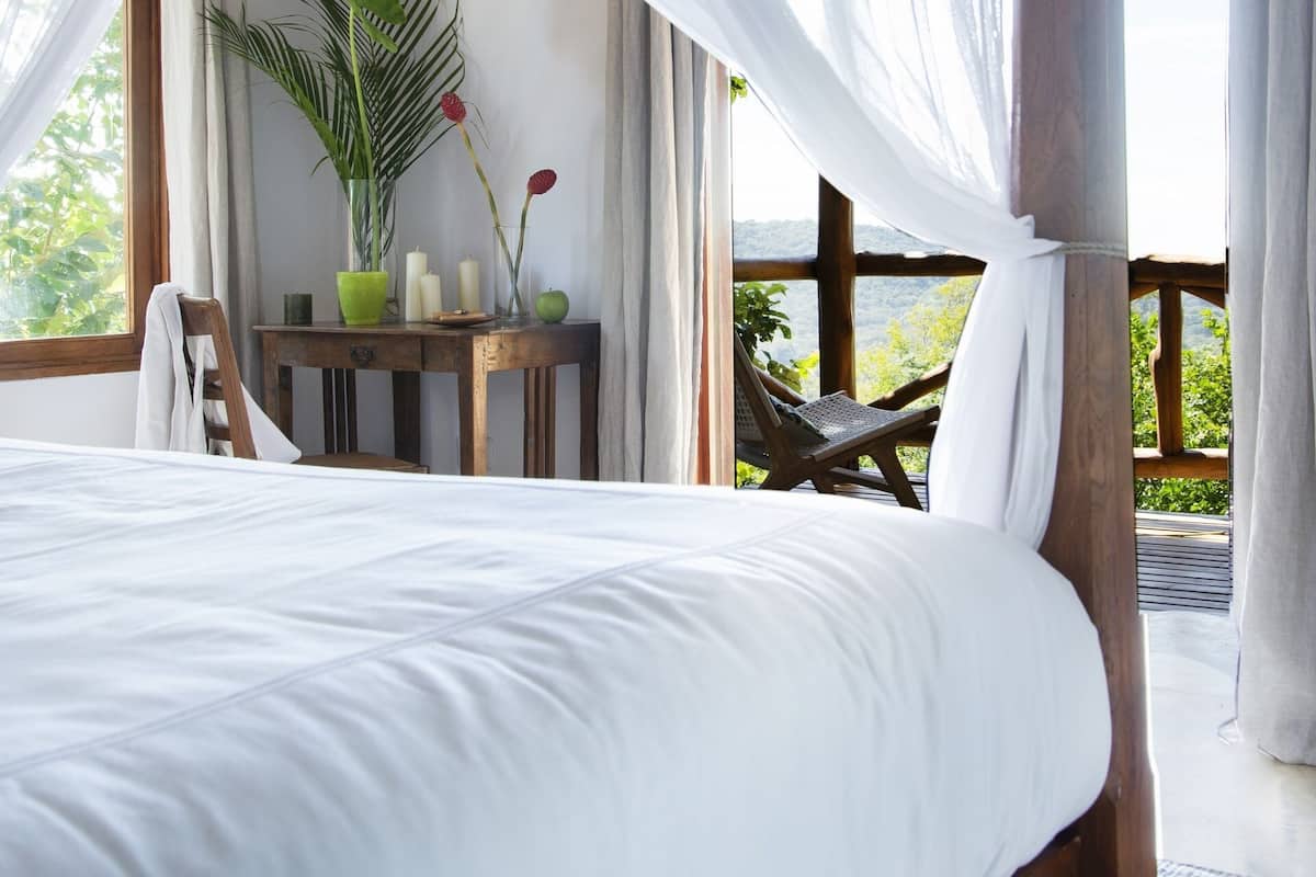 Ein Schlafzimmer mit Baldachinbett, einem Holztisch mit Kerzen, einer Topfpflanze und Blick ins Freie.