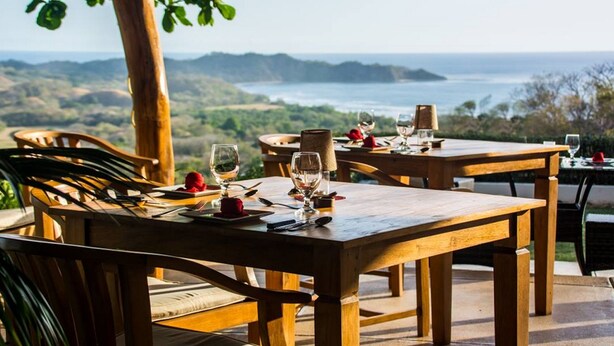 A wooden dining table set with plates, cutlery, and glasses, overlooking a scenic landscape with hills and a body of water.