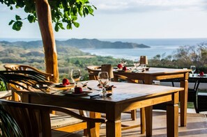 A wooden dining table set with plates, cutlery, and glasses, overlooking a scenic landscape with hills and a body of water. - Tierra Magnifica Boutique Hotel (Nosara)