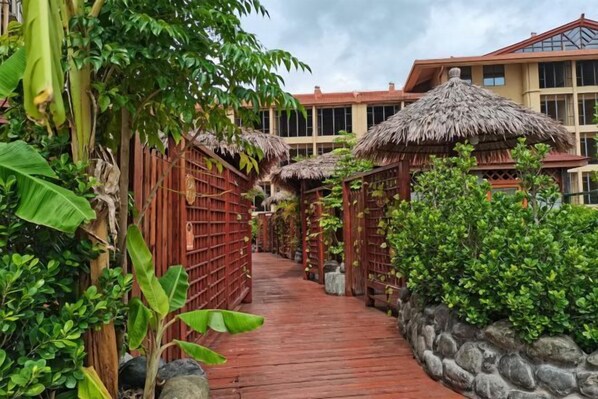 Un sendero de madera que conduce a un edificio con construcciones de techo de paja y abundante vegetación.