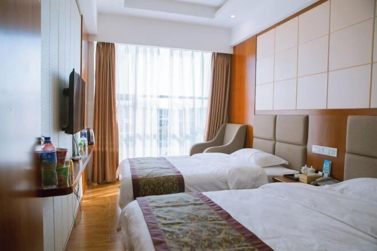 Habitación de hotel con dos camas, un televisor, repisas con artículos de aseo y un ventanal con cortinas.