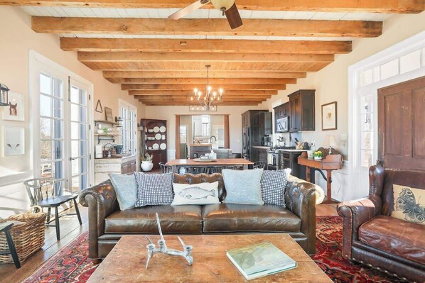 A spacious living room with a leather sofa, wooden ceiling beams, and a chandelier.