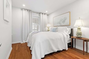 A bedroom with a bed, two bedside lamps, a window with curtains, a framed artwork on the wall, and a wooden floor.