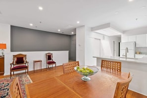A modern kitchen with white cabinets, a dining area with a wooden table and chairs, and a fruit bowl on the table.
