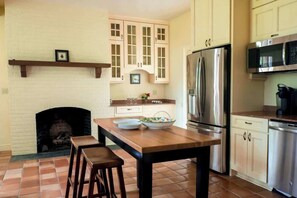 A kitchen with a fireplace, wooden cabinets, a stainless steel refrigerator, and a wooden dining table with stools. - Hoos` Homestead | Historic UVA area home (Charlottesville)