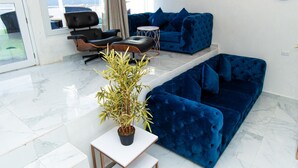 Un salon moderne avec un canapé en velours bleu, un fauteuil en cuir noir et une petite table ronde. Une plante en pot est posée sur une table blanche, et une grande fenêtre laisse entrer la lumière naturelle.
