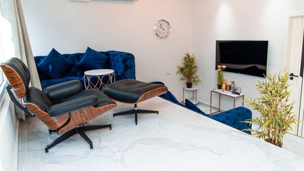 Un salon moderne comprenant un canapé, deux fauteuils, une petite table ronde, une plante en pot et un téléviseur fixé au mur.