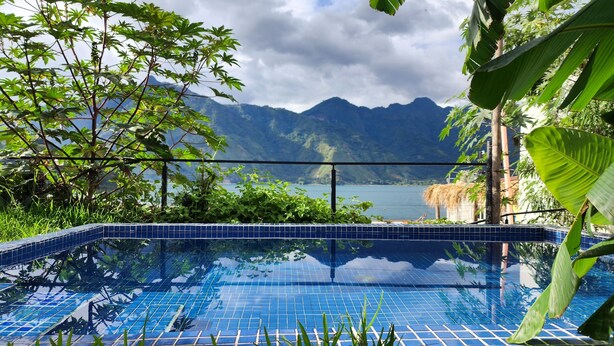 Una piscina azul cristalina con bordes de azulejos, rodeada de abundante vegetación y con montañas al fondo.