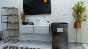 Una sala de estar moderna con un televisor de pantalla plana montado en la pared, un refrigerador pequeño, una consola blanca con adornos, un estante de mimbre y una planta en maceta.