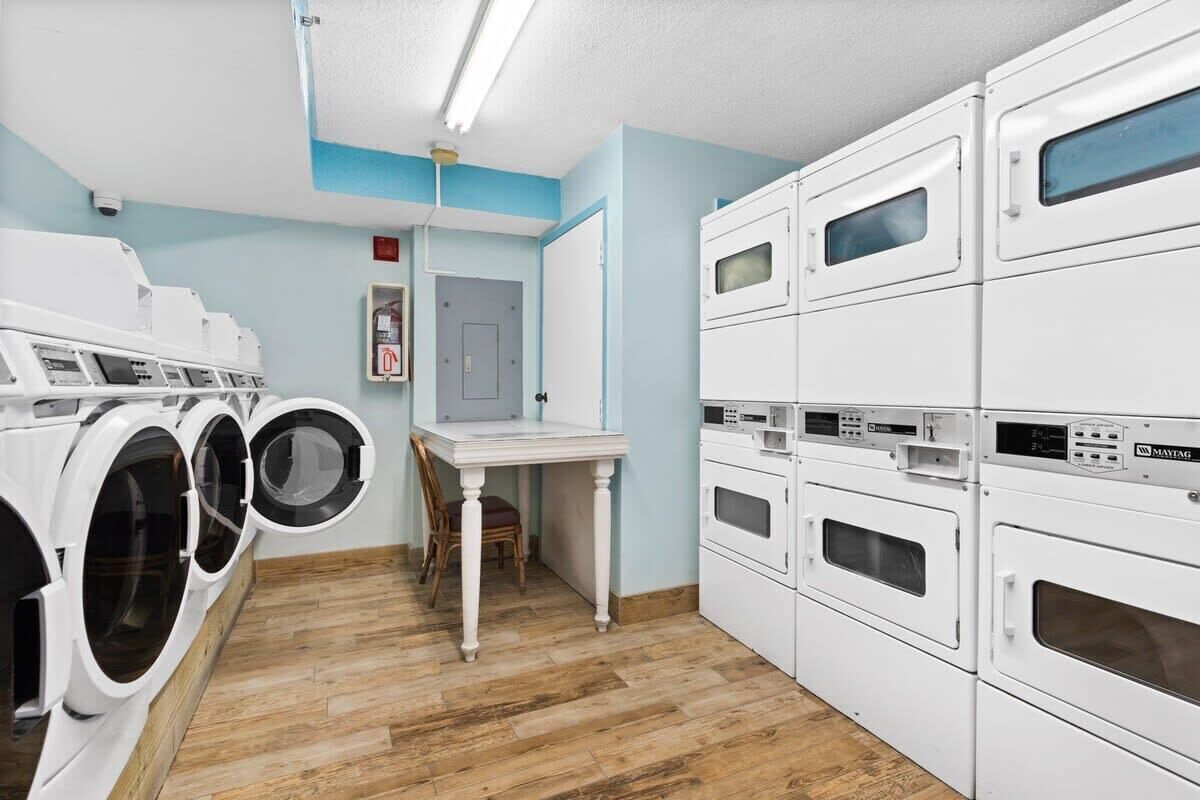 A laundry room with multiple washing machines and dryers, a small table, and a chair.