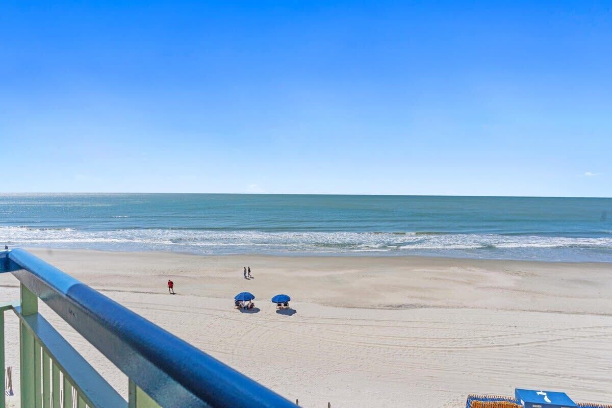 Vista a la playa desde un balcón con barandilla, una playa de arena y el océano.